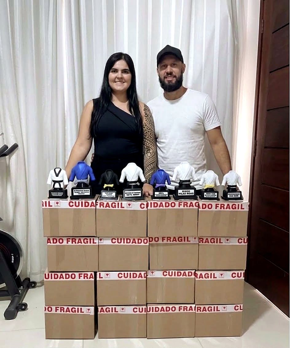 O casal proprietário da THP3D ao lado de vários troféus prontos sobre caixas de envio com fita de cuidado frágil